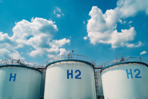 large hydrogen storage tanks. "H2" written on it, indicating it stores hydrogen gas. The tanks are white, equipped with ladders and railings, and are set against a blue sky with scattered clouds.