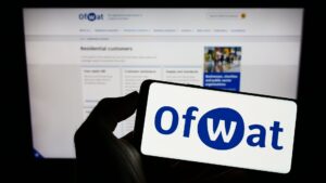 Person holding cellphone with logo of Water Services Regulation Authority (Ofwat) on screen in front of webpage. Focus on phone display.